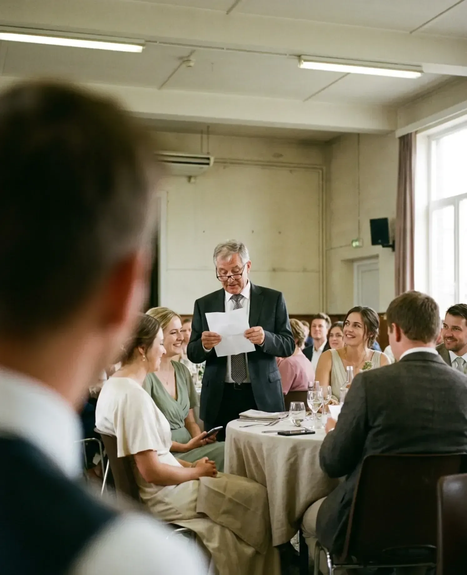 Een man leest een speech voor van een blad papier, gezien vanaf een stoel tussen de gasten.